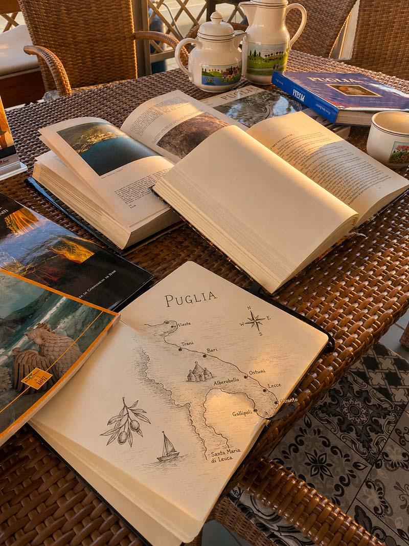 Scrivania di ricerca: libri di storia barese, taccuino aperto con appunti su Bari Vecchia, cartoline d’epoca della città, mappa storica e quotidiano ‘L’Eco di Bari’ sulle reliquie di San Nicola